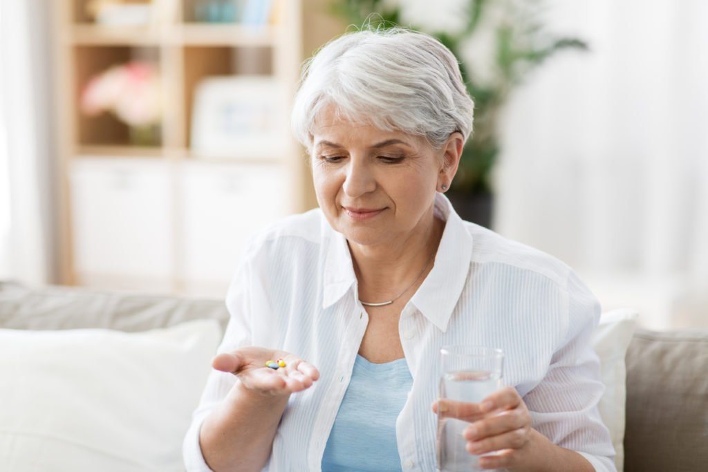 A mature woman prepares to take her medication held in one hand, a glass of water in the other, sitting on her couch. 