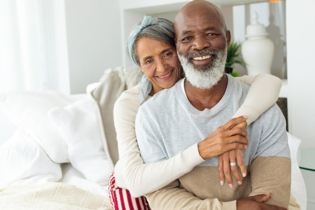 A mature woman wraps her arms around her mature husband on the couch, both smiling. 
