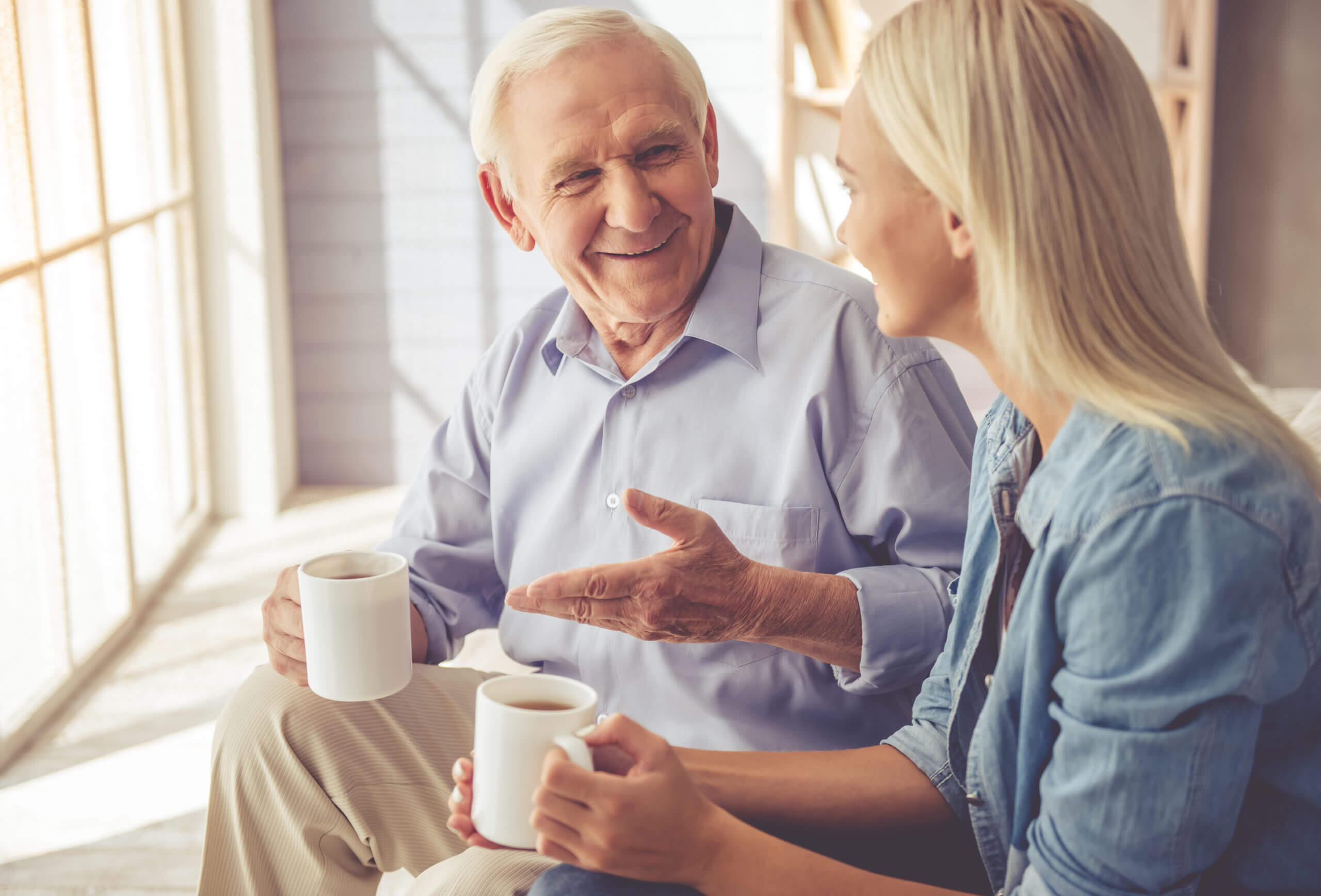A mature man and his young female caregiver enjoy coffee together by an open, sunny window.