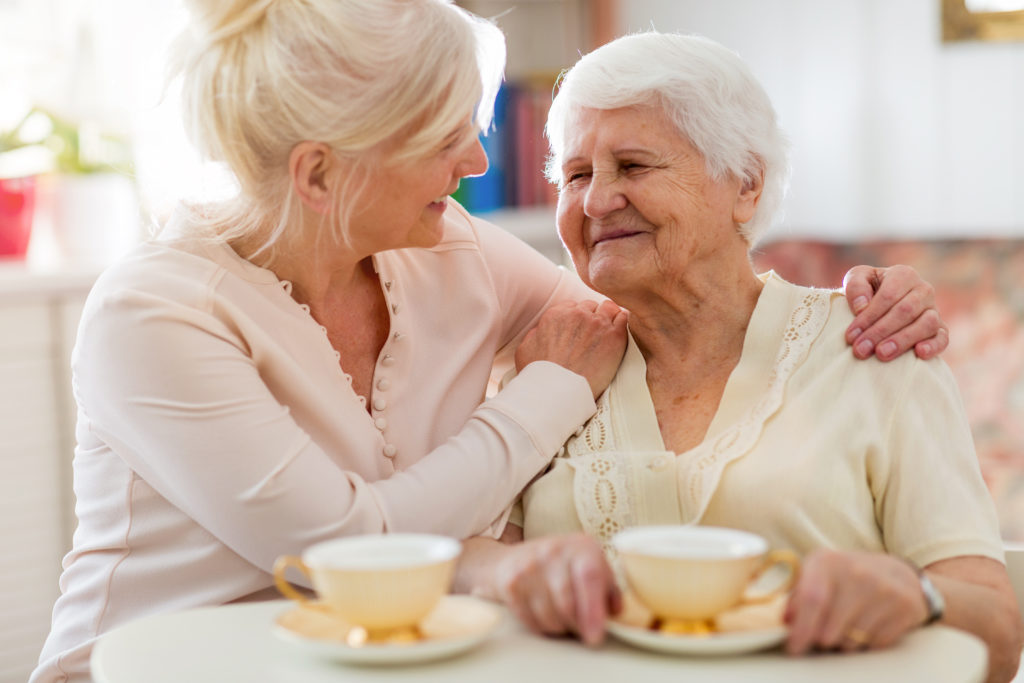 Smiling older woman holds a cup of tea while her daughter embraces her. 
