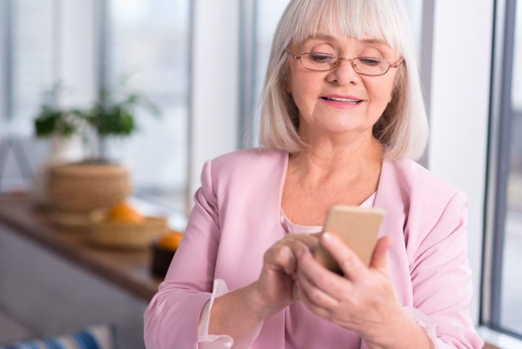 Senior woman with white hair and glasses looks at a smartphone while sending a message. 