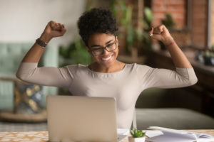 A confident black woman wearing glasses raises her arms in triumph from good news on her laptop.