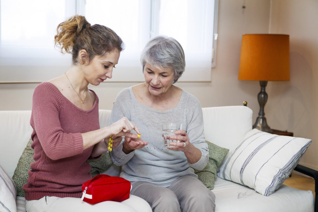 A female adult daughter helps her elderly mother with water glass take prescribed medication, both seated on couch.