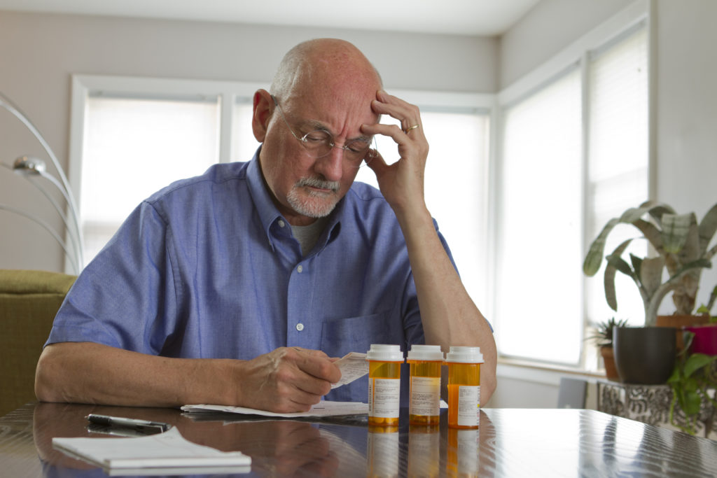 A senior man wearing glasses holds his temple in concern, prescribed medication in front of him on table. 
