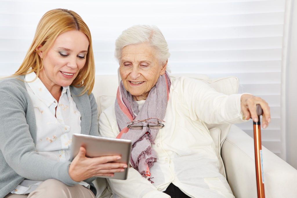 A middle-aged woman shows an older female senior with a cane how to use assisted living technology on a tablet. 