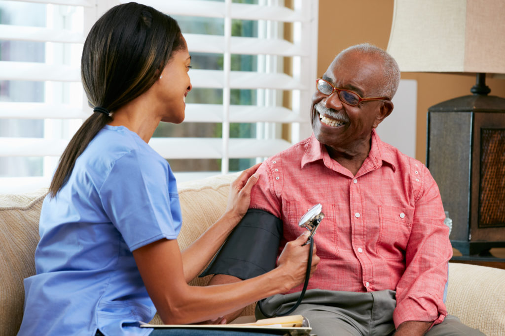 Happy Black senior care nurse takes the blood pressure of Black male resident also smiling.
