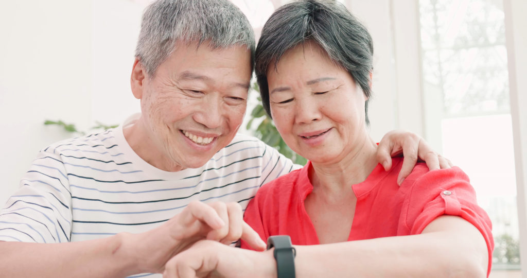 Smiling Asian senior couple embrace, with husband pointing to wife’s smartwatch.