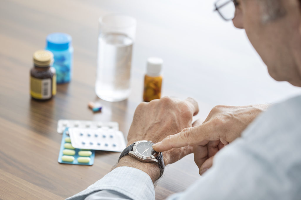 Male senior points to his wristwatch, indicating time to take nearby medications with glass of water.
