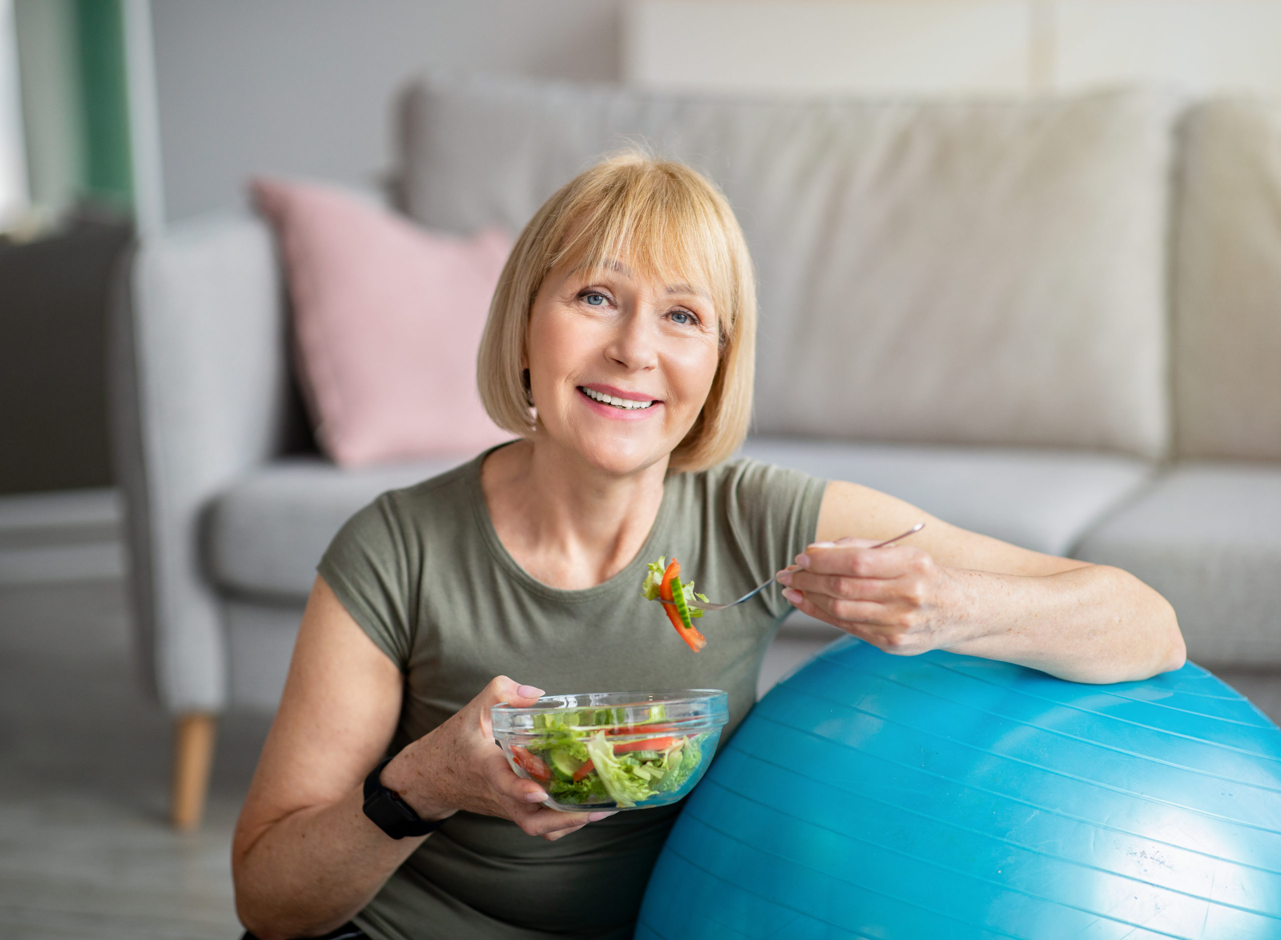  A senior woman eats a salad and smiles while seated next to an exercise ball.