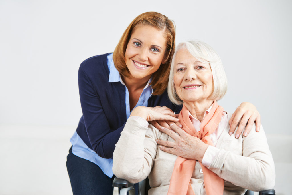 Elderly woman seated, hugging younger female caregiver while both smile at the camera.