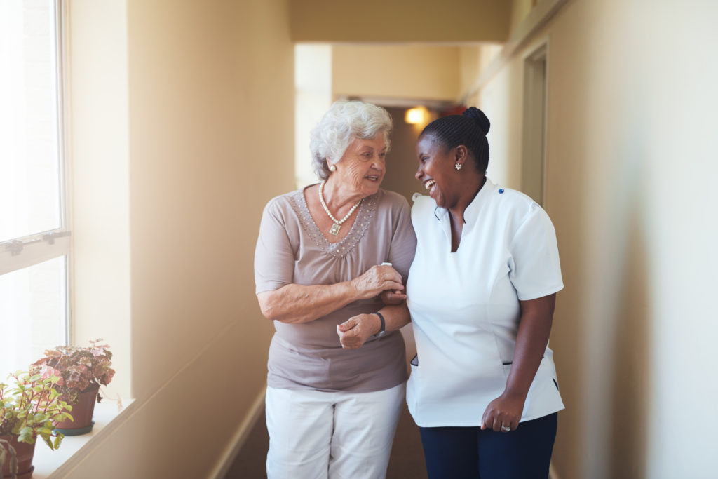 Portrait of happy female caregiver and senior woman walking down a hall in a senior living community. 