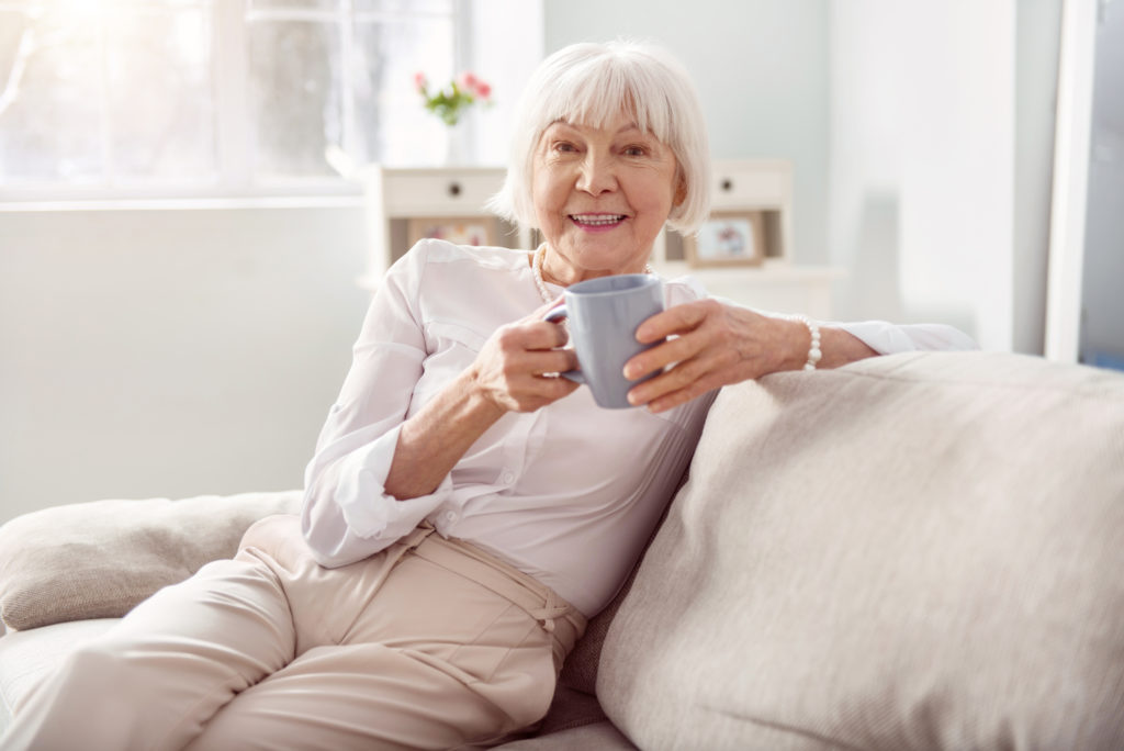 Smiling older woman enjoys a drink in a mug on the sofa. 