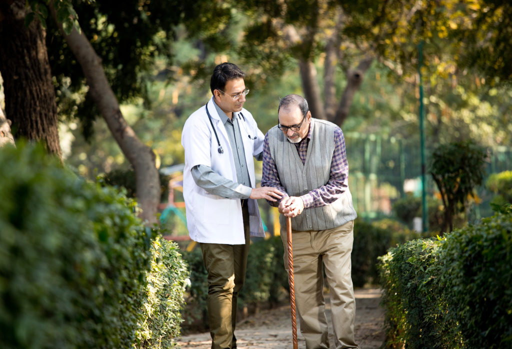 A doctor helps a man walk with a cane in a garden park. 