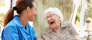 Woman wearing a senior care technology bracelet sits outside on sunny day, laughing with her smiling professional caregiver.