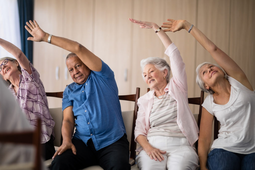  Four senior living community residents sit in chairs, stretching their left arms above their heads during an exercise class.