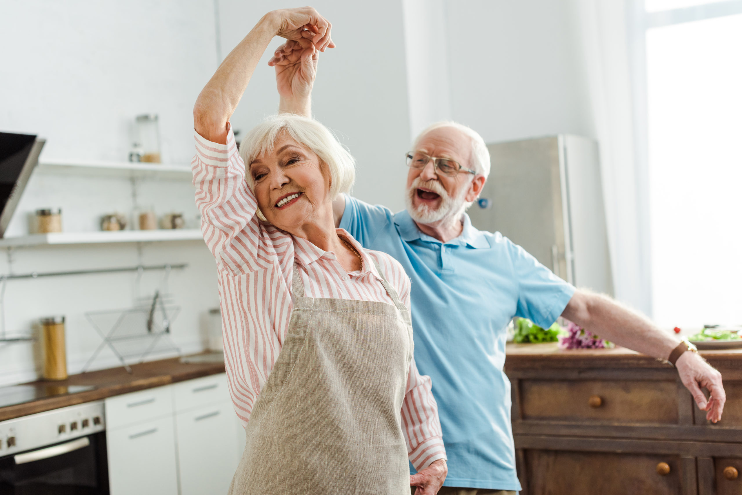 Happy senior couple dances together in their kitchen. 