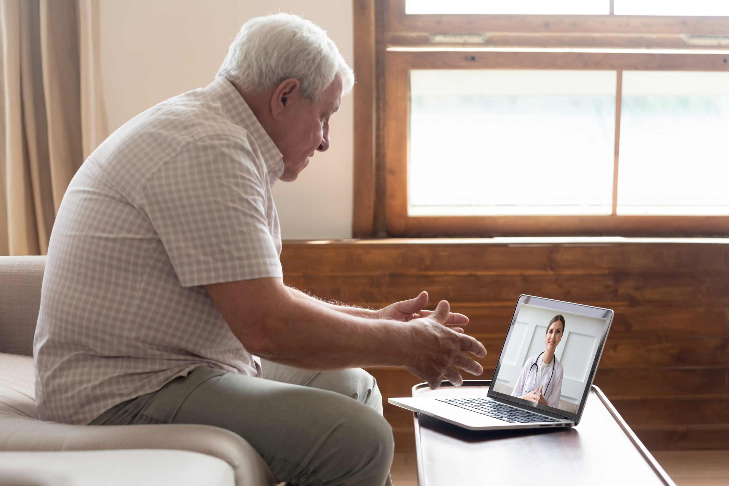 A senior man sits on the couch and talks to his doctor via a laptop. 