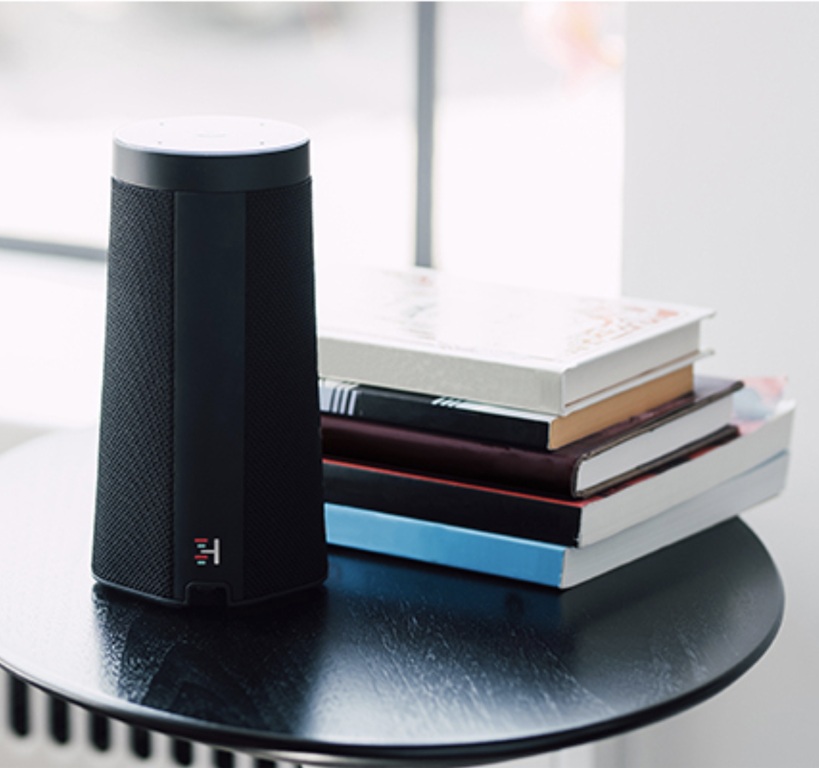 WellBe smart speaker on a side table next to a stack of books. 
