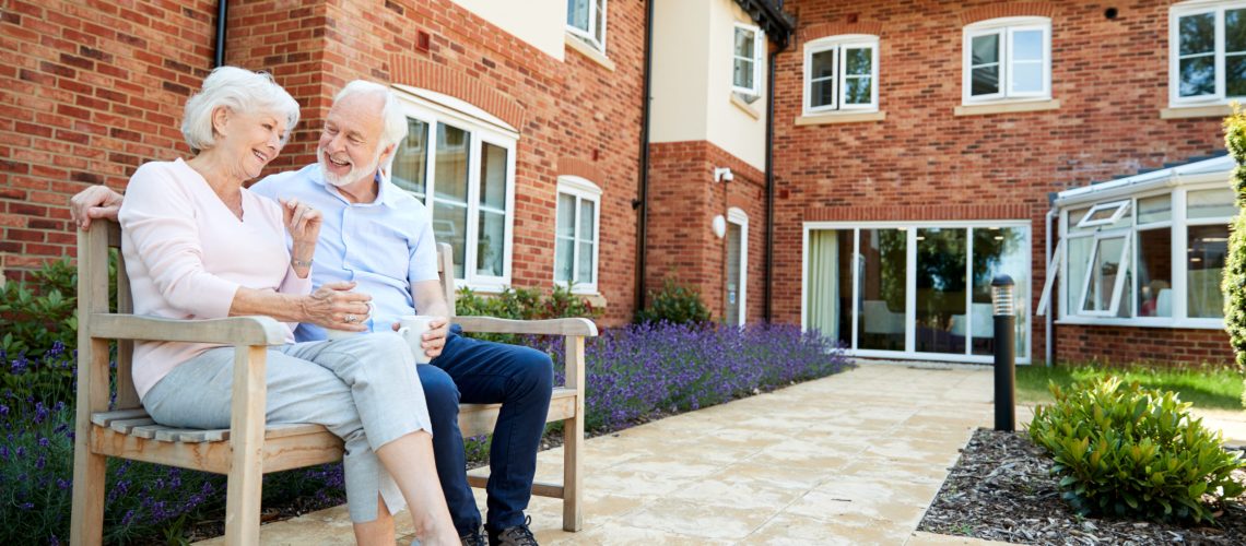 Senior couple sits on a bench outside of their beautiful brick assisted-living building.