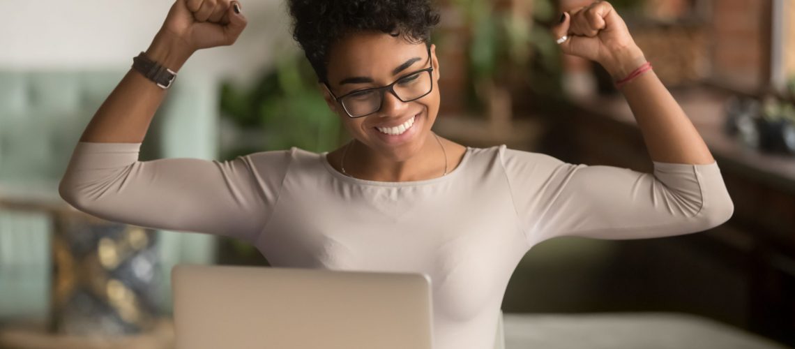 A confident black woman wearing glasses raises her arms in triumph from good news on her laptop.
