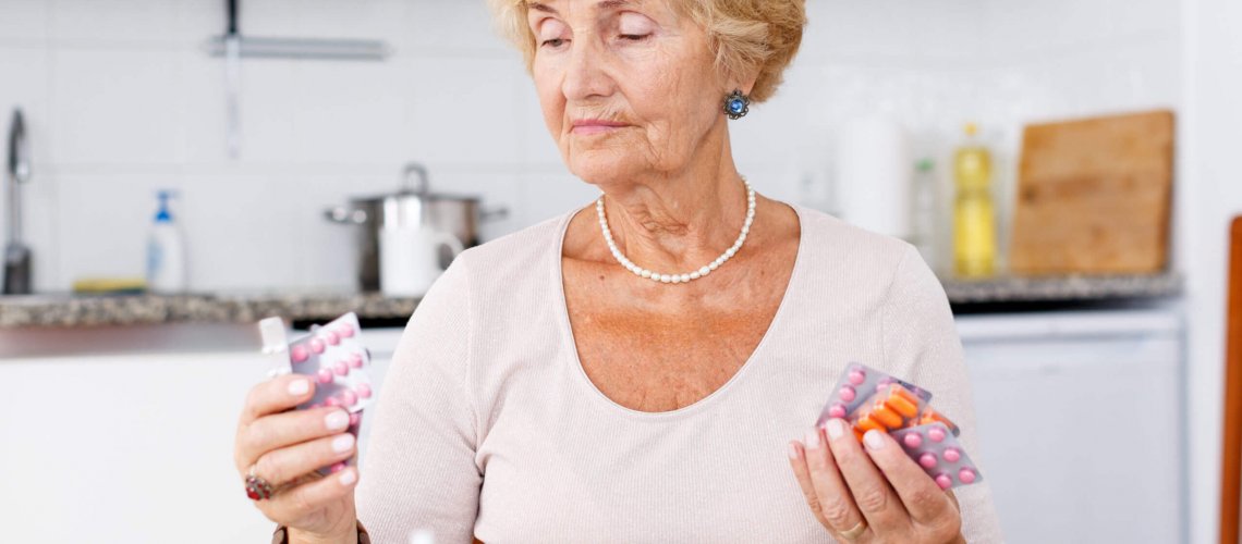 A senior woman holds multiple packs of prescribed medication having difficulty with medication management.