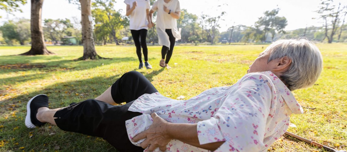 Senior woman holding her hip lying on the grass with a cane on the ground beside her.