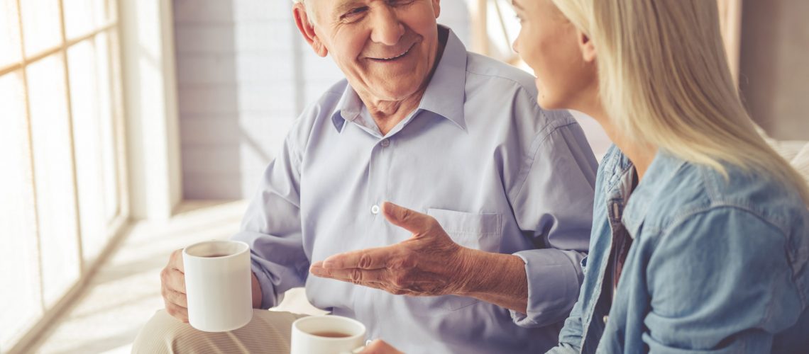A mature man and his young female caregiver enjoy coffee together by an open, sunny window.